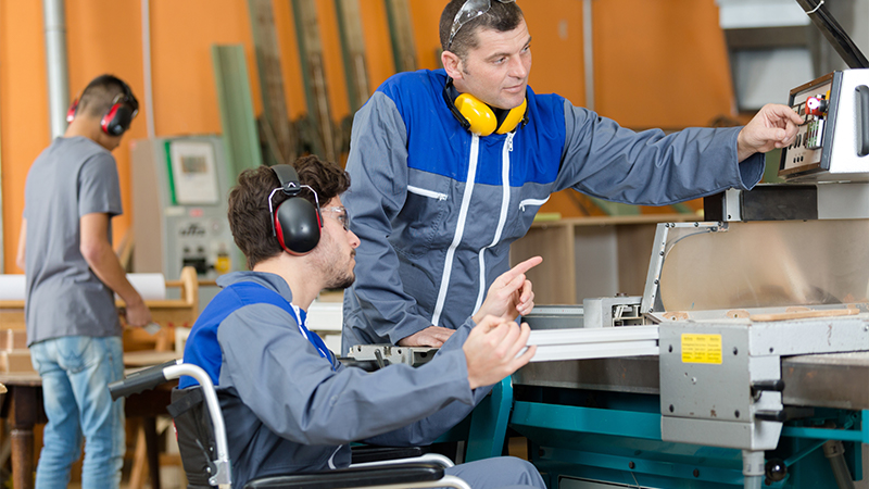 3 Personen mit Gehörschutz in einer Werkhalle. eine Person davon im Rollstuhl, wird von einer zweiten Person eingewiesen.